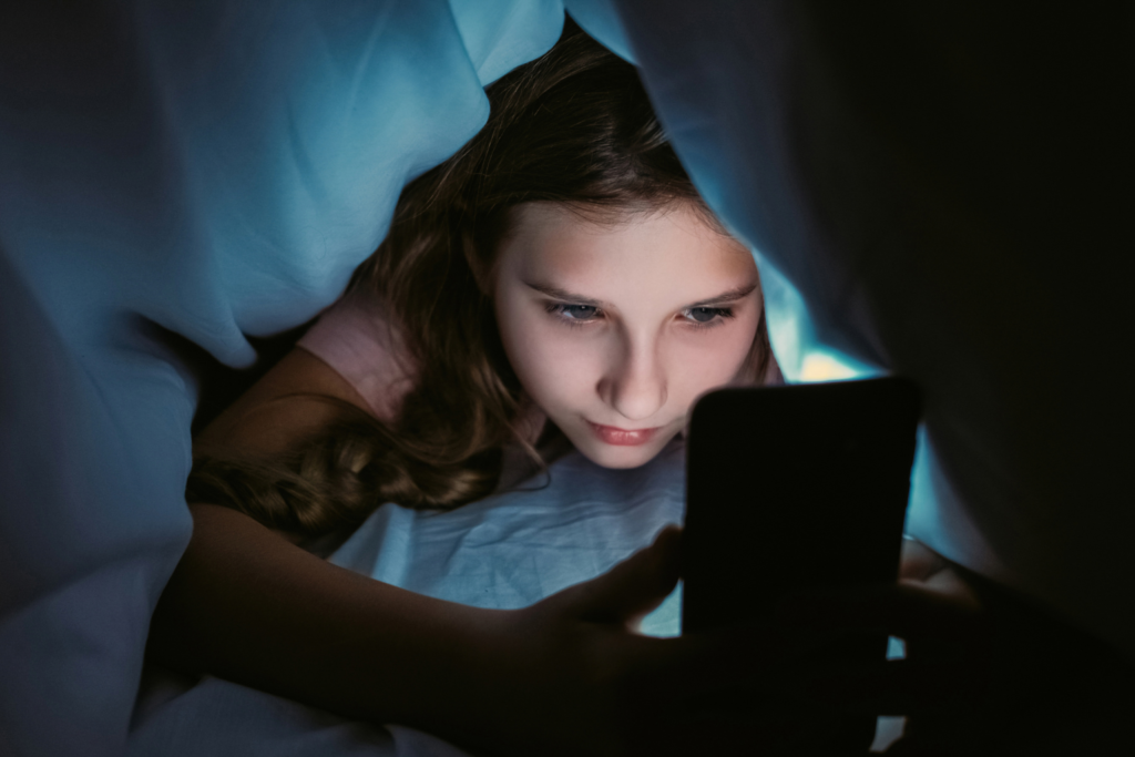 Smutna nastolatka leżąca pod kołdrą i wpatrzona w ekran smartfona w nocy, symbolizująca cyfrową izolację i samotność pokolenia Z.