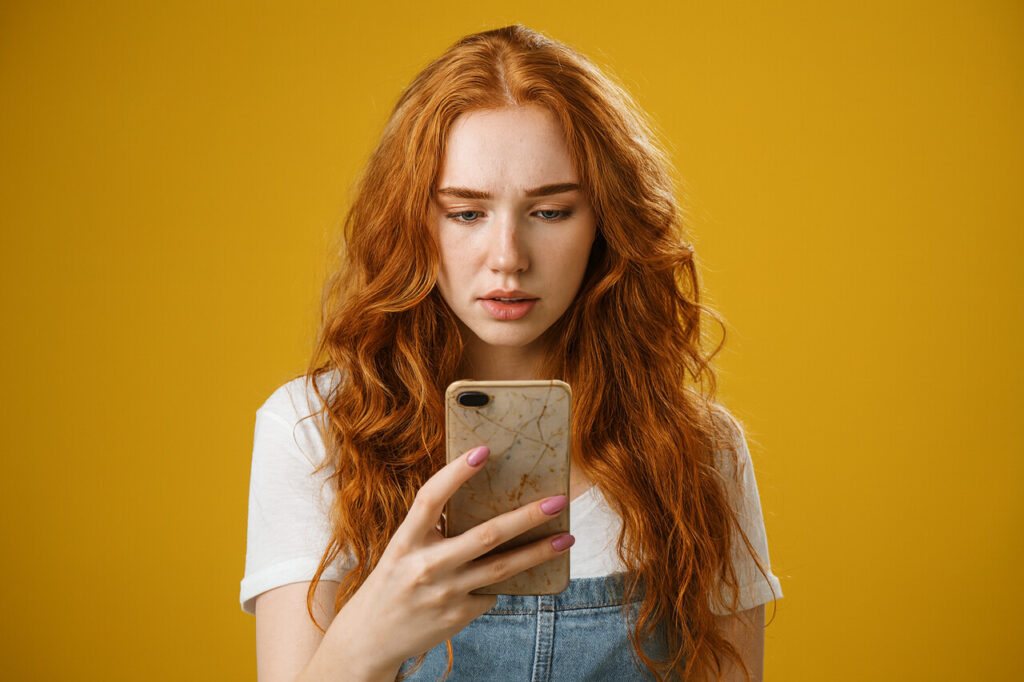 Zaniepokojona młoda kobieta o rudych włosach patrzy na ekran smartfona, szukając numeru telefonu zaufania lub natychmiastowej pomocy psychologicznej w sytuacji kryzysu emocjonalnego.